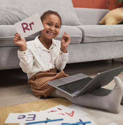 Picture of a girl sitting on the floor, a laptop on her legs, holding up a card with the letter R on it